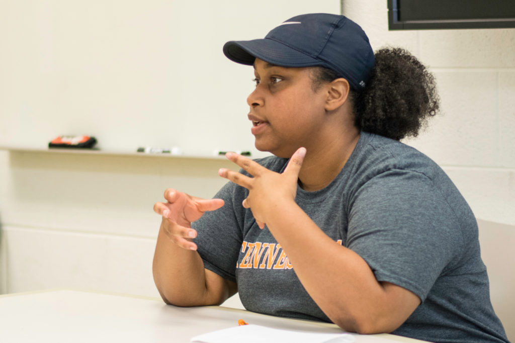 a student in baseball hat and t shirt speaks as they sit at a table in a classroom 