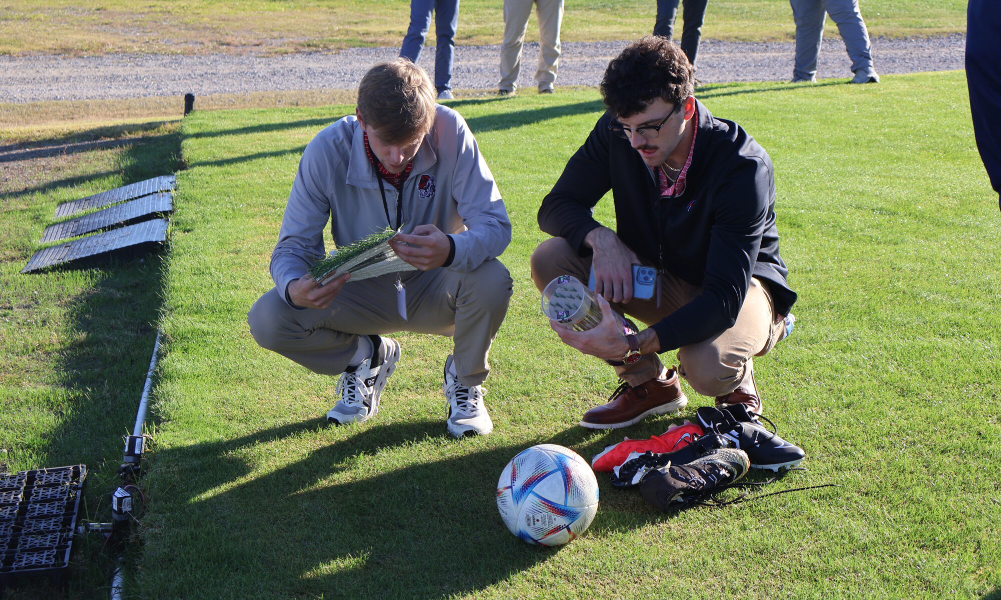 Two men kneeling down on grass looking at a ball, grass samples, and shoes