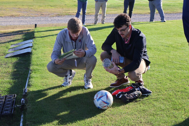 Two men kneeling down on grass looking at a ball, grass samples, and shoes