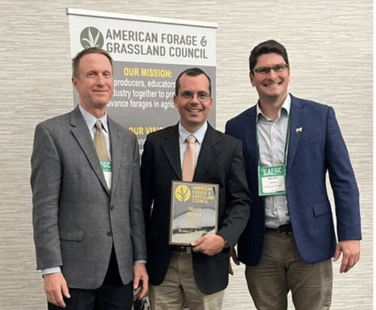 Three men smiling with the man in the middle holding an award
