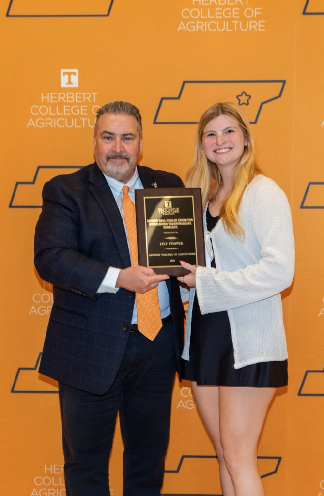 Photo of a woman and a man holding an award in front of an orange backdrop