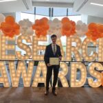 Student standing in front of light up letters that read "RESEARCH AWARDS" with orange and white balloons, holding an award