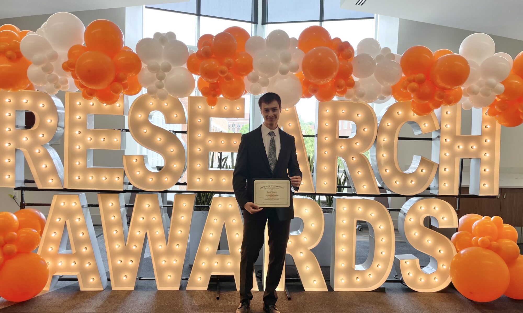 Student standing in front of light up letters that read "RESEARCH AWARDS" with orange and white balloons, holding an award