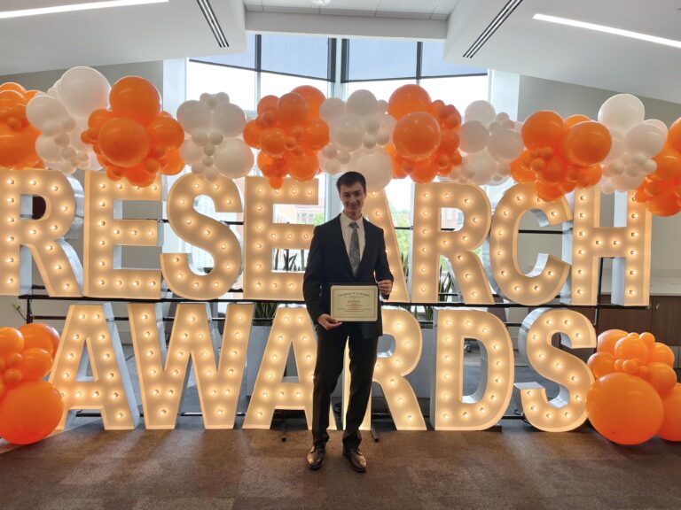 Student standing in front of light up letters that read "RESEARCH AWARDS" with orange and white balloons, holding an award