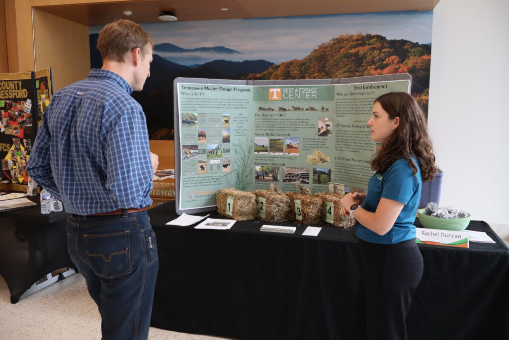 Photo of a woman talking to a man in front of a table with a poster board and bales of hay on it