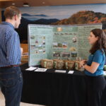 Photo of a woman talking to a man in front of a table with a poster board and bales of hay on it