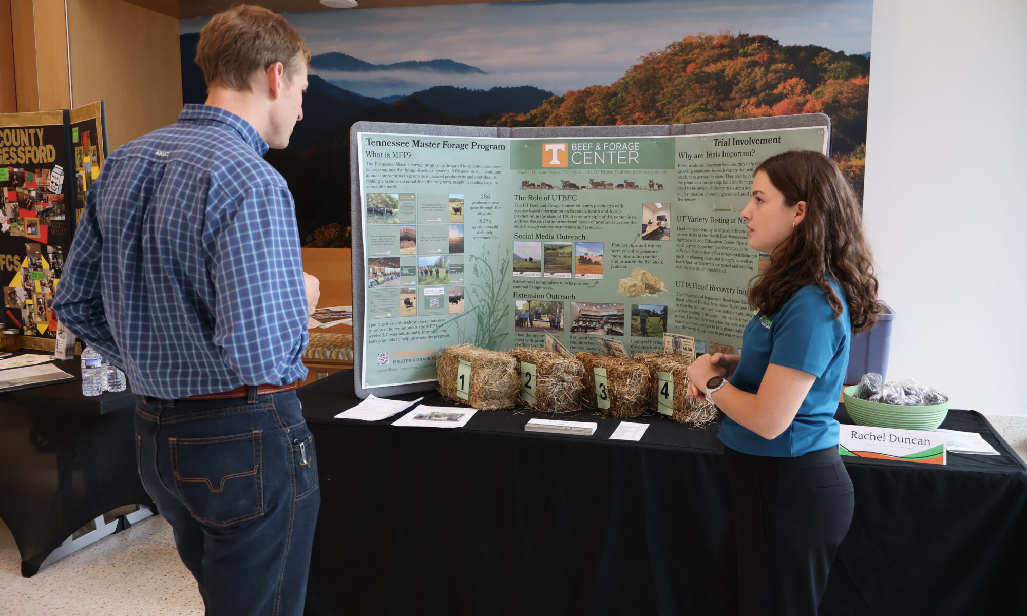 Photo of a woman talking to a man in front of a table with a poster board and bales of hay on it