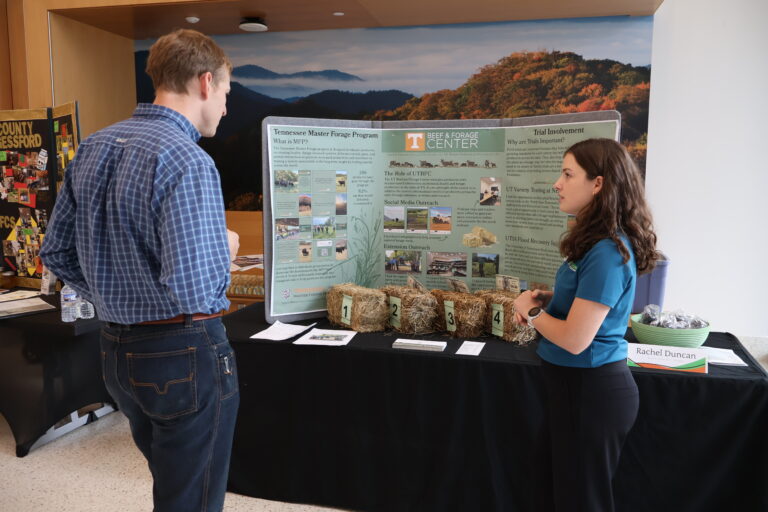 Photo of a woman talking to a man in front of a table with a poster board and bales of hay on it