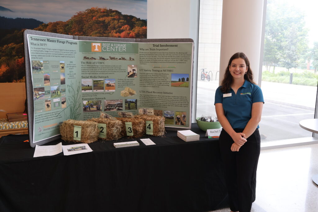 Photo of a woman standing in front of a table with a poster board and bales of hay on it