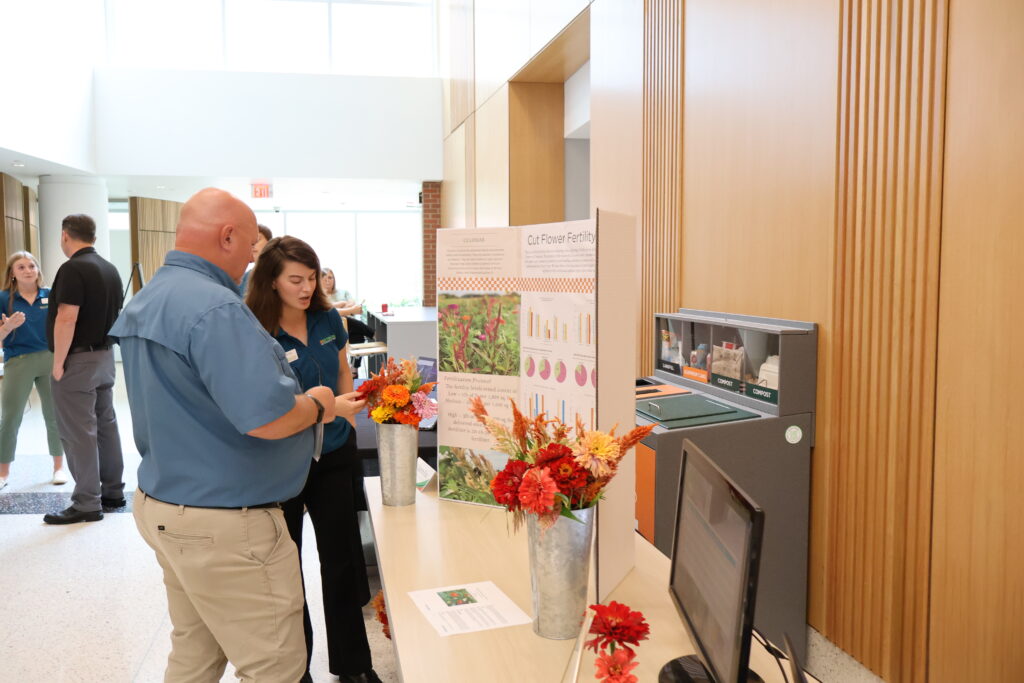 Photo of a woman talking to a man in front of a booth with a poster board, computer monitor, and flowers in vases on it