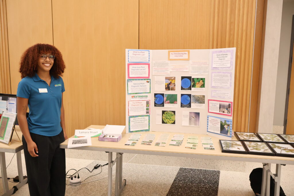 Photo of a woman standing in front of a table display with a poster board, rack cards, display insects, and framed plants