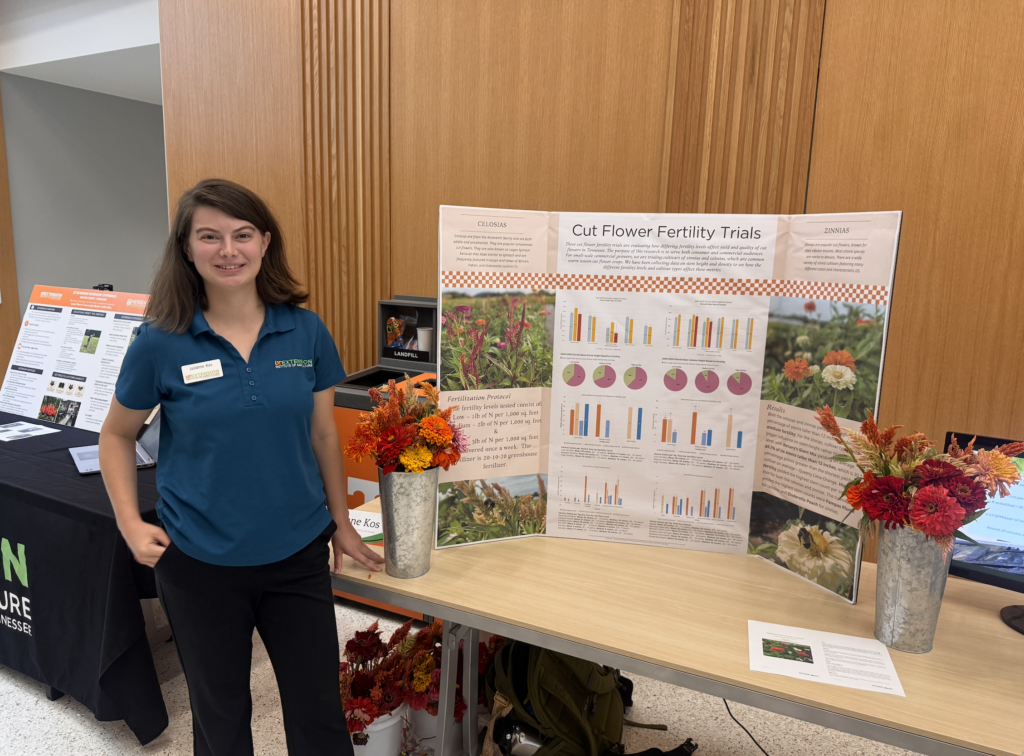 Phot of a woman standing in front of a table with two vases of flowers, a computer monitor, and a poster board